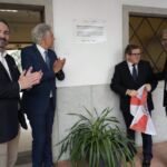 Quatro homens junto a uma placa de um centro tecnológico especializado; dois aplaudem, enquanto outro segura uma bandeira. A imagem sugere uma cerimónia de inauguração, mas a placa não permite confirmar a data nem os participantes.
