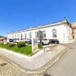 Vista exterior de um edifício branco de grandes dimensões, com janelas em arco, junto a uma praça pavimentada. Em frente há carros estacionados e uma zona ajardinada.