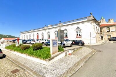 Vista exterior de um edifício branco de grandes dimensões, com janelas em arco, junto a uma praça pavimentada. Em frente há carros estacionados e uma zona ajardinada.