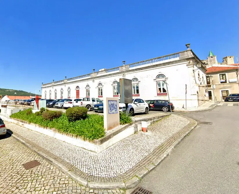 Vista exterior de um edifício branco de grandes dimensões, com janelas em arco, junto a uma praça pavimentada. Em frente há carros estacionados e uma zona ajardinada.