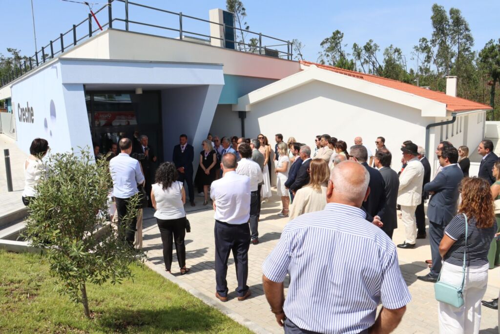 Um grupo de pessoas está reunido à entrada de um edifício com a palavra «Creche» na fachada. Algumas parecem ouvir uma intervenção junto à porta.