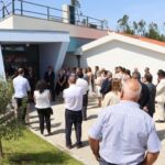 Um grupo de pessoas está reunido à entrada de um edifício com a palavra «Creche» na fachada. Algumas parecem ouvir uma intervenção junto à porta.