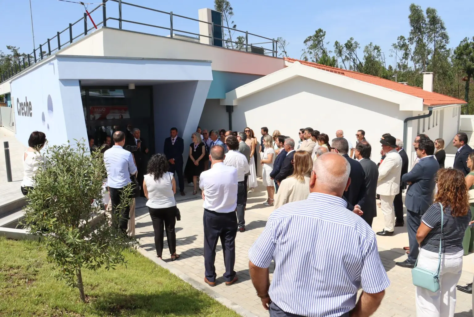 Um grupo de pessoas está reunido à entrada de um edifício com a palavra «Creche» na fachada. Algumas parecem ouvir uma intervenção junto à porta.