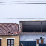 A imagem mostra fachadas de edifícios contíguos, com telhados de telha e cabos aéreos. No edifício da direita, vê-se uma estrutura elevada e uma vedação escura.