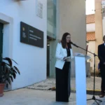 Uma mulher discursa ao microfone, atrás de um púlpito, à entrada de um edifício. Um homem de fato acompanha a intervenção.
