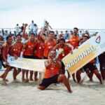 Uma equipa de andebol de praia celebra na areia, perante uma bancada com público. Os jogadores exibem cartazes com as palavras «Campeão» e «Nacional», associados à Federação de Andebol de Portugal.