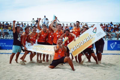 Uma equipa de andebol de praia celebra na areia, perante uma bancada com público. Os jogadores exibem cartazes com as palavras «Campeão» e «Nacional», associados à Federação de Andebol de Portugal.