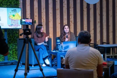 Duas mulheres participam numa conversa perante o público, com microfones e um computador portátil. Um ecrã apresenta um cartaz do HA HA Art Film Festival.