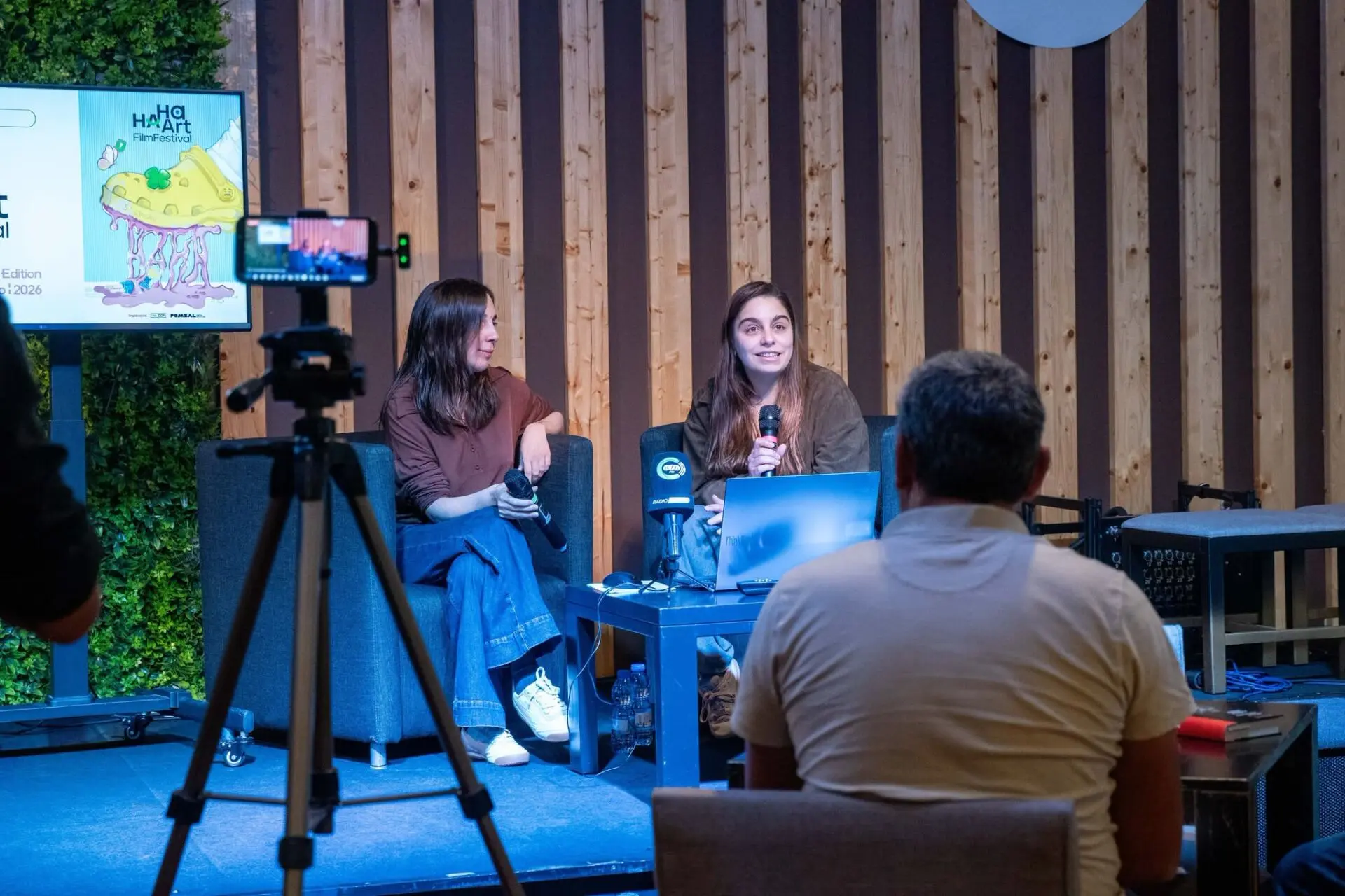 Duas mulheres participam numa conversa perante o público, com microfones e um computador portátil. Um ecrã apresenta um cartaz do HA HA Art Film Festival.