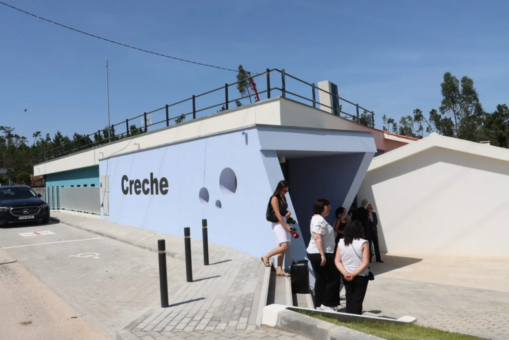 Exterior de um edifício com a palavra «Creche» na fachada. Várias pessoas encontram-se junto à entrada e há lugares de estacionamento à frente.