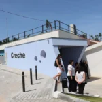 Exterior de um edifício com a palavra «Creche» na fachada. Várias pessoas encontram-se junto à entrada e há lugares de estacionamento à frente.