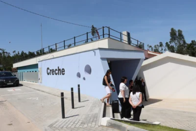 Exterior de um edifício com a palavra «Creche» na fachada. Várias pessoas encontram-se junto à entrada e há lugares de estacionamento à frente.