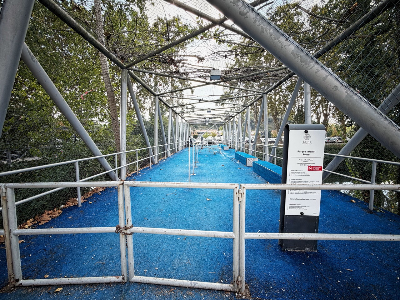 Um portão fechado impede o acesso a um parque infantil com piso azul e estrutura de rede. A placa à entrada identifica-o como «Parque Infantil Ponte», em Leiria.