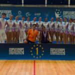 Um grupo de patinadores artísticos posa com um troféu junto a um painel com o número 1. A imagem mostra a inscrição «Cup of Europe Figueres 2026» num pavilhão desportivo.