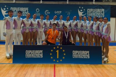Um grupo de patinadores artísticos posa com um troféu junto a um painel com o número 1. A imagem mostra a inscrição «Cup of Europe Figueres 2026» num pavilhão desportivo.