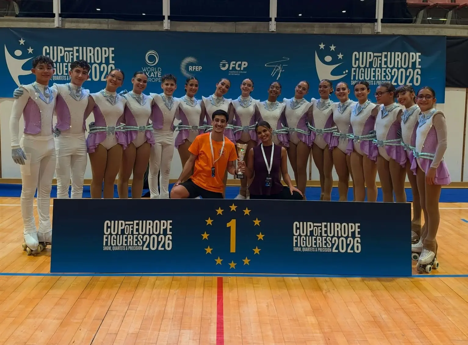 Um grupo de patinadores artísticos posa com um troféu junto a um painel com o número 1. A imagem mostra a inscrição «Cup of Europe Figueres 2026» num pavilhão desportivo.