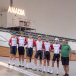 Seis ciclistas com equipamentos de Portugal e um homem de polo verde posam numa pista de ciclismo coberta. Ao fundo, vê-se um letreiro luminoso com a palavra «ANADIA».