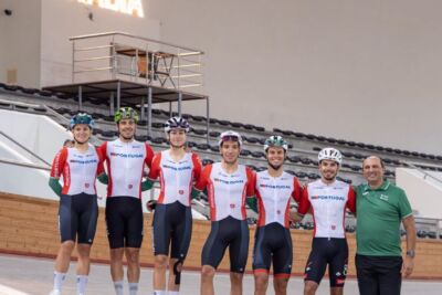 Seis ciclistas com equipamentos de Portugal e um homem de polo verde posam numa pista de ciclismo coberta. Ao fundo, vê-se um letreiro luminoso com a palavra «ANADIA».