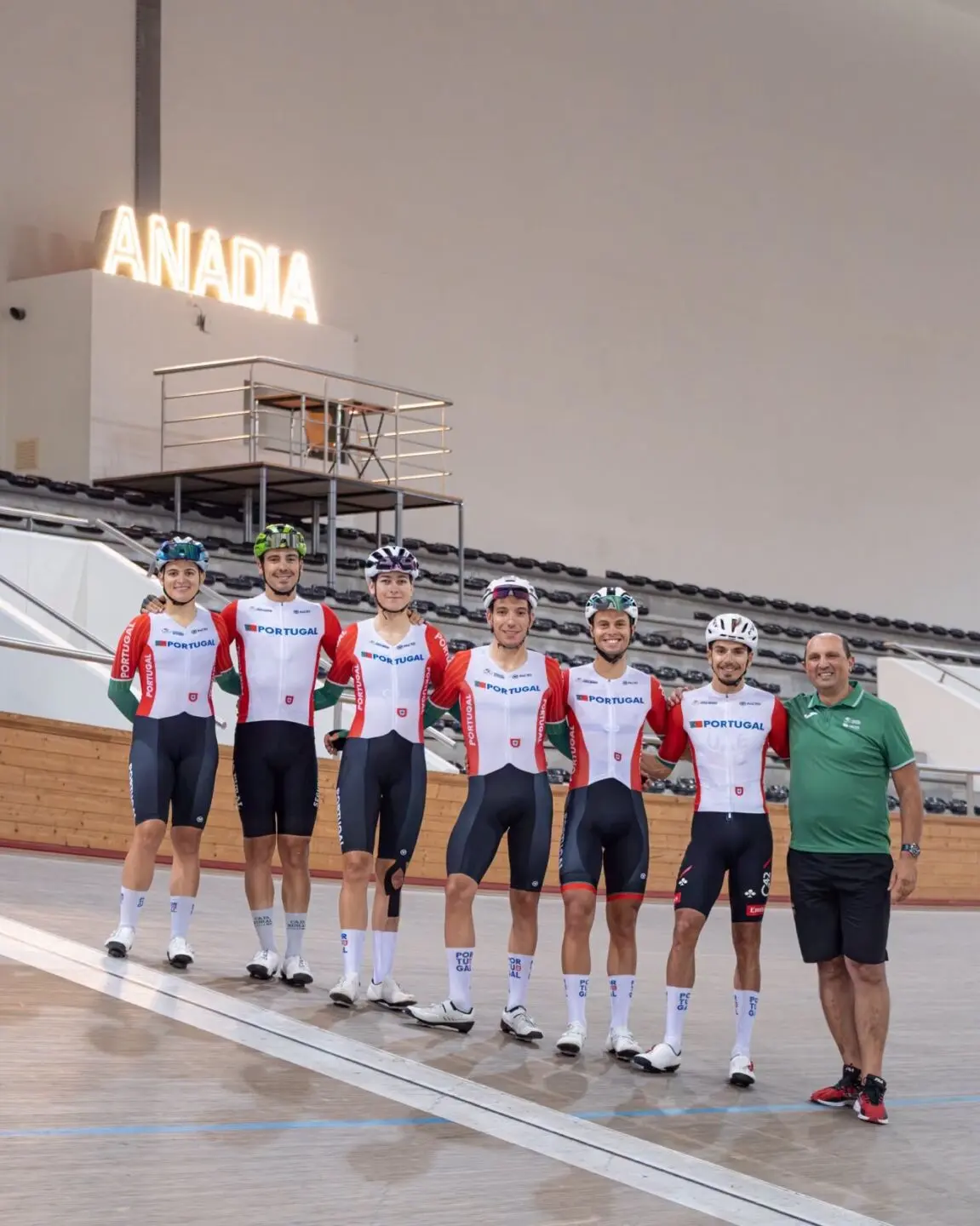 Seis ciclistas com equipamentos de Portugal e um homem de polo verde posam numa pista de ciclismo coberta. Ao fundo, vê-se um letreiro luminoso com a palavra «ANADIA».