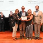 Um grupo posa em palco com um certificado, enquanto outras pessoas aplaudem. No ecrã ao fundo são visíveis referências a prémios de comunicações orais e posters e a um encontro nacional.