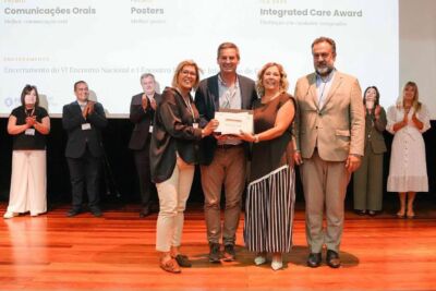 Um grupo posa em palco com um certificado, enquanto outras pessoas aplaudem. No ecrã ao fundo são visíveis referências a prémios de comunicações orais e posters e a um encontro nacional.