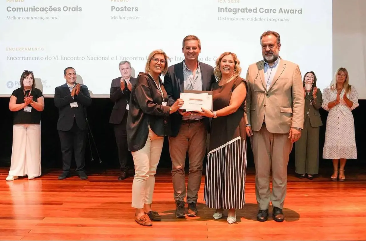 Um grupo posa em palco com um certificado, enquanto outras pessoas aplaudem. No ecrã ao fundo são visíveis referências a prémios de comunicações orais e posters e a um encontro nacional.