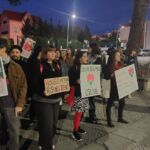 Um grupo de pessoas manifesta-se ao anoitecer, segurando cartazes que reivindicam o direito à habitação e o cumprimento do artigo 65.º da Constituição. Num dos cartazes lê-se «Casa para todos» e «Cumprir Abril».