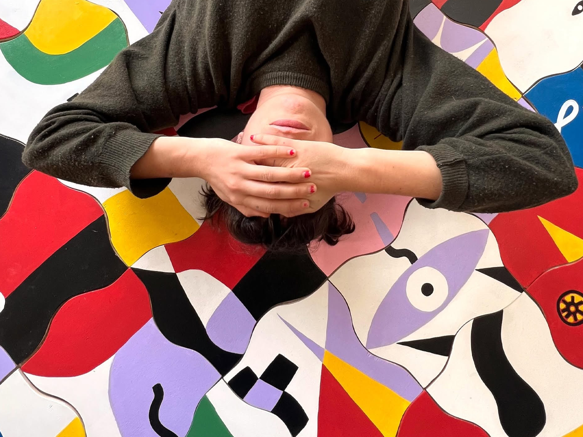 Uma pessoa deitada cobre os olhos com as mãos sobre uma superfície ilustrada com formas abstratas e cores vivas.