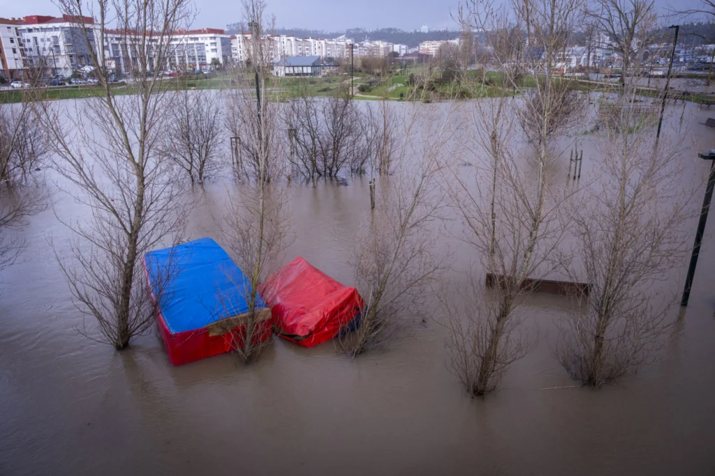 Uma zona verde urbana está inundada, com árvores e estruturas cobertas por lonas parcialmente submersas. Veem-se edifícios residenciais ao fundo.