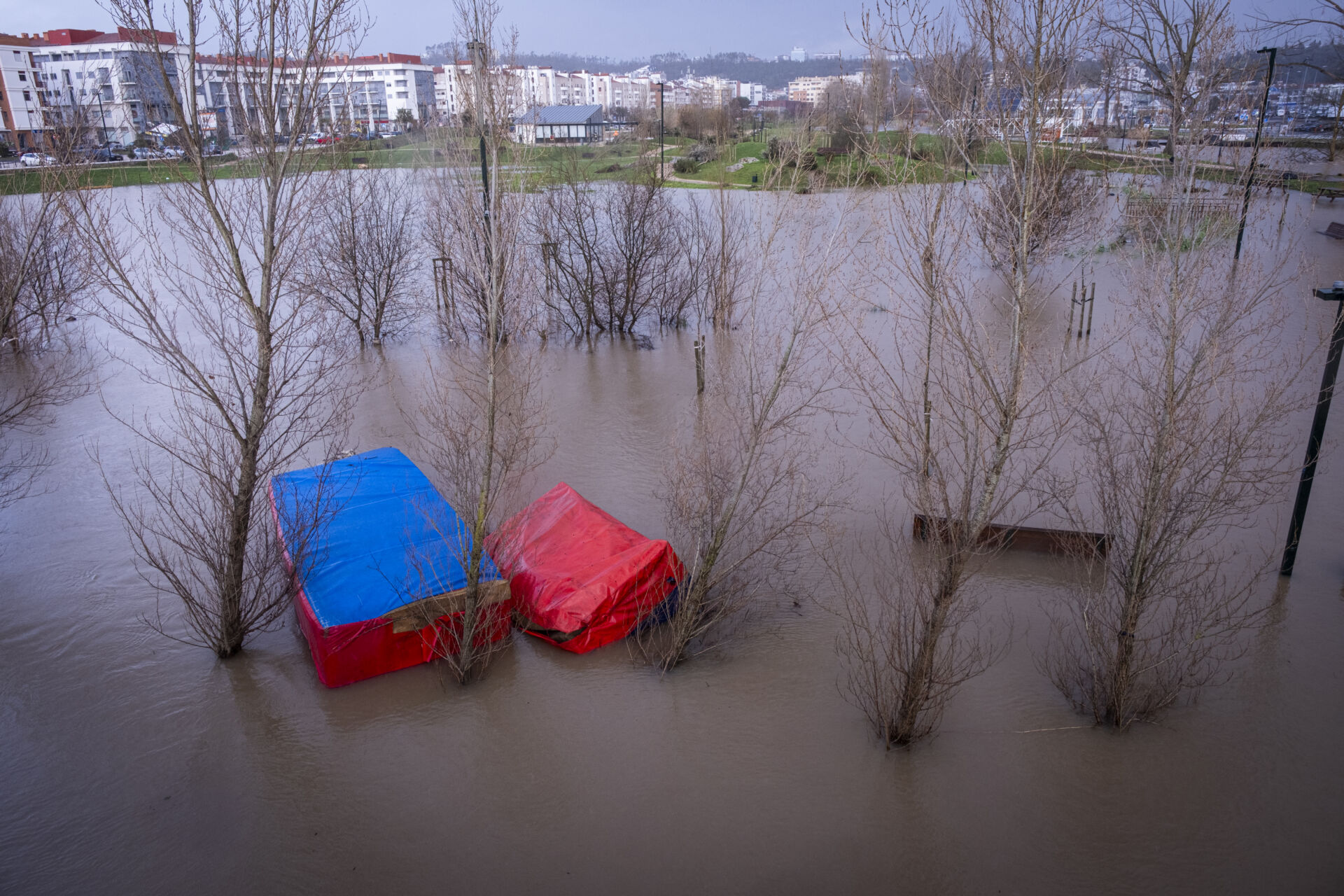 Uma zona verde urbana está inundada, com árvores e estruturas cobertas por lonas parcialmente submersas. Veem-se edifícios residenciais ao fundo.