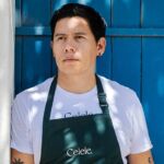 Retrato de uma pessoa com uma t-shirt branca e um avental escuro com a inscrição «Celele.», junto a uma porta azul. Não há texto visível que permita identificar a pessoa.