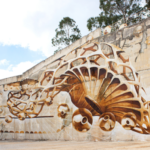 Um mural de arte urbana ocupa uma grande parede de betão ao ar livre. A composição abstrata, em tons castanhos e brancos, combina formas orgânicas, linhas e círculos.