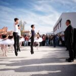 Um grupo de jovens apresenta uma dança de ballet ao ar livre, diante de espectadores e de homens de fato. A atuação decorre junto a um edifício moderno.