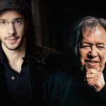 Retrato de dois homens lado a lado, diante de um fundo escuro. O homem à esquerda usa boné e óculos; o da direita apoia o queixo na mão.