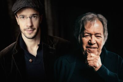 Retrato de dois homens lado a lado, diante de um fundo escuro. O homem à esquerda usa boné e óculos; o da direita apoia o queixo na mão.