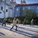 Um grupo de pessoas pratica movimentos coordenados ao ar livre, numa praça junto a edifícios históricos. A luz baixa projeta sombras longas no pavimento.