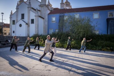 Um grupo de pessoas pratica movimentos coordenados ao ar livre, numa praça junto a edifícios históricos. A luz baixa projeta sombras longas no pavimento.