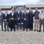 Um grupo de pessoas posa no exterior, diante de tratores e de uma viatura amarela. Algumas vestem uniformes de serviço e outras usam fatos.