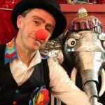 Um homem com chapéu preto e nariz vermelho de palhaço posa ao lado de uma figura de elefante feita de peças metálicas. O fundo tem tecidos vermelhos.
