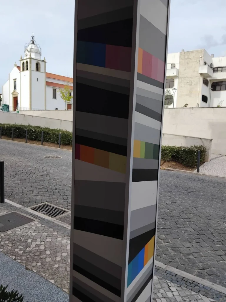 Uma coluna de arte urbana com faixas geométricas e coloridas ocupa o primeiro plano. Ao fundo, vê-se uma igreja branca junto a uma rua empedrada.