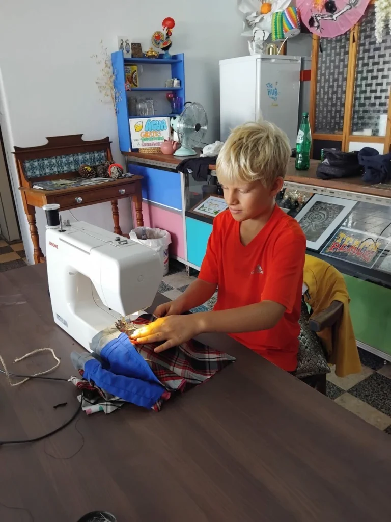 Uma pessoa jovem costura tecidos numa máquina de costura, sentada à mesa num espaço com vários objetos e peças de roupa. Ao fundo, vê-se um cartaz sobre água grátis.