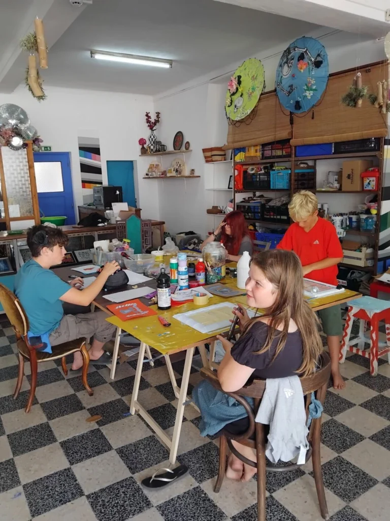 Quatro jovens estão reunidos à volta de uma mesa com tintas, pincéis e outros materiais num espaço de trabalhos manuais. Nas paredes veem-se peças decorativas coloridas.