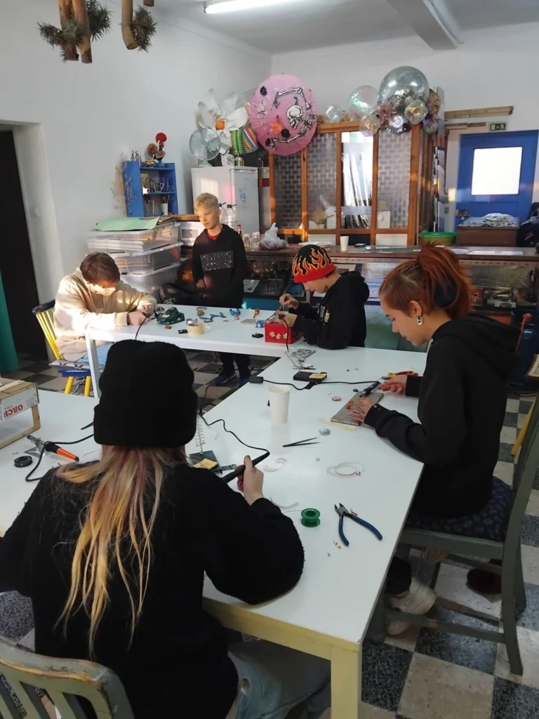 Cinco pessoas estão reunidas à volta de mesas, a trabalhar com pequenos objetos e ferramentas num espaço de oficina. Veem-se alicates, fios e materiais de artesanato.