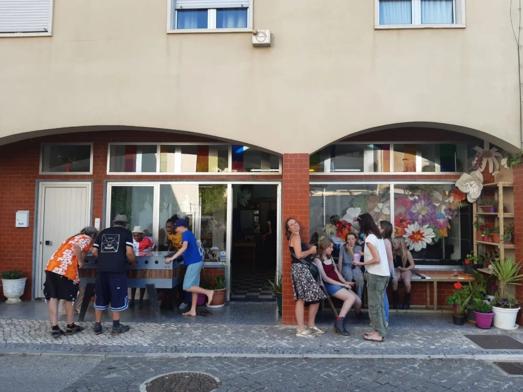 Um grupo de pessoas convive à porta de um edifício, junto a uma mesa de matraquilhos. A montra está decorada com flores de papel e há vasos com plantas à entrada.