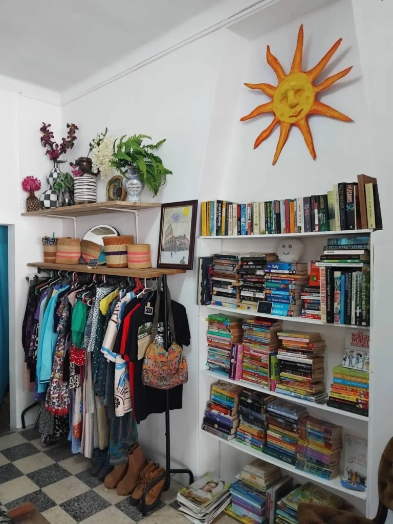 Interior de uma loja com livros em estantes e roupa em cabides. Há vasos, cestos e uma decoração em forma de sol na parede.