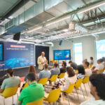 Um orador apresenta uma palestra perante uma plateia sentada. No palco lê-se «PC DIG4 STAGE» e «Empowering the Future»; os ecrãs mostram «From Vision to Scale».